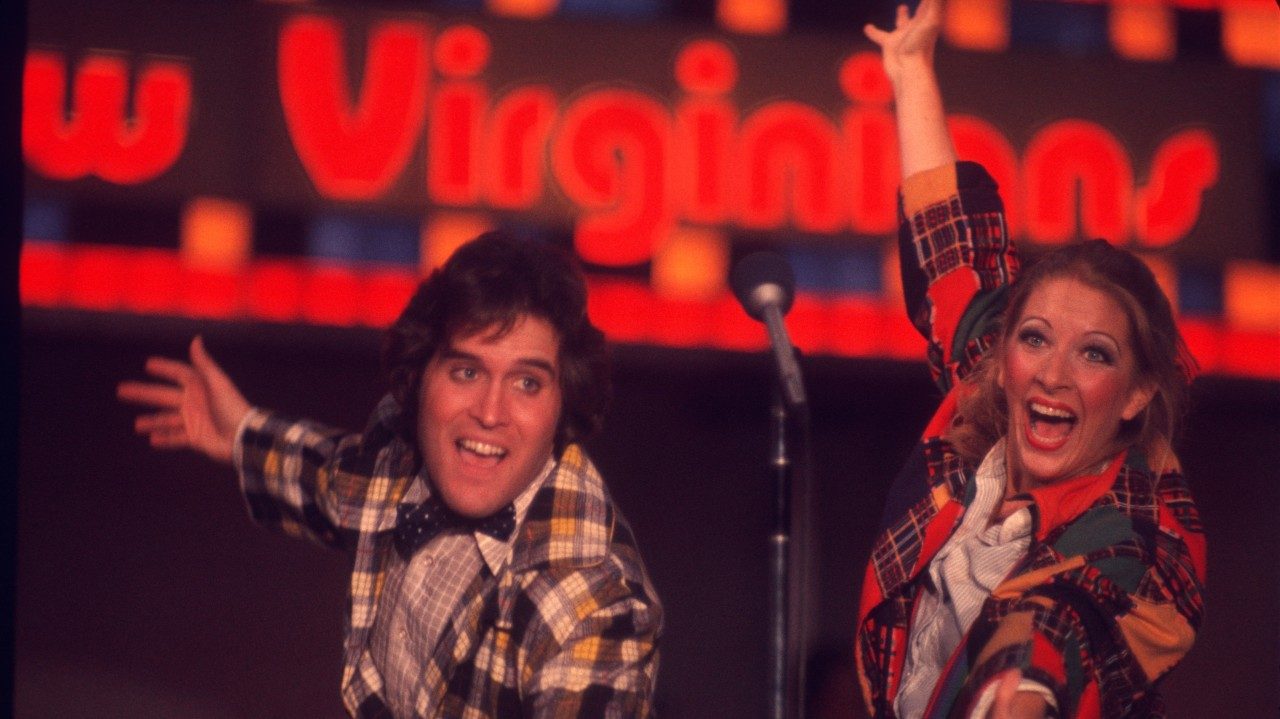  Two members of the New Virginians perform on stage. They sing into a shared microphone on a stand, leaning their left arms towards the audience. The man, on left, is a white man with medium length dark brown hair, and the woman, on the right, is a white woman with long blonde hair, a full face of makeup, and thin brown eyebrows. The both wear plaid multicolor jackets.
