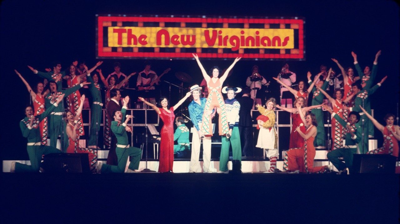  The New Virginians perform on stage at the end of a song or show. Everyone has one hand raised high above their heads. In the middle, two men support a woman on their shoulders. She wears and orange jumpsuit and raises both of her arms above her head. Behind them all is a light up sign that reads "The New Virginians."