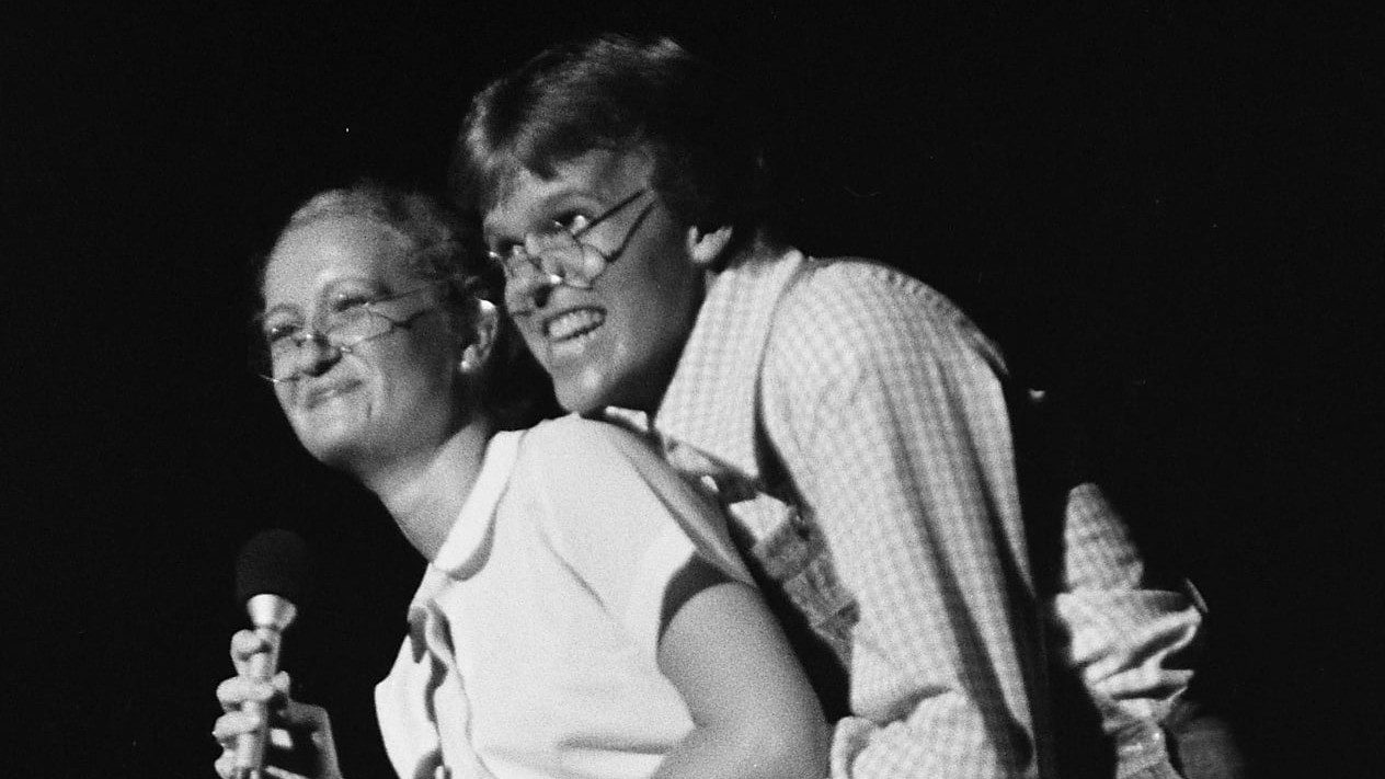 Two members of the New Virginians sing and perform on stage in this black and white image. On the left/in front is a white woman with blonde or light colored hair in a white button down short sleeved shirt and glasses. On the right/behind is a white man with medium length brown hair, plaid button down shirt, and glasses. He hugs her from behind. Both are smiling.