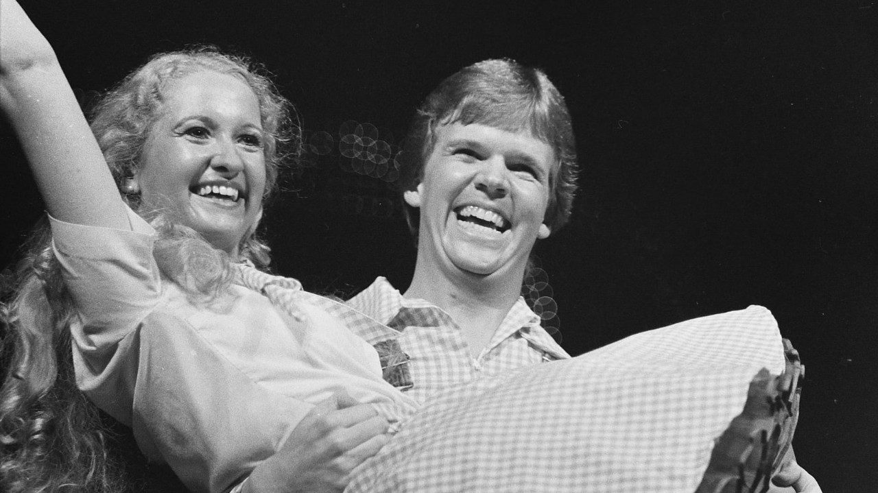  Two members of the New Virginians perform on stage in this black and white image. White man with medium length light hair smiles and holds a woman in his arms. She is a white woman with long, wavy light hair, wearing a costume with a white button down short sleeved top and plaid checkered skirt with matching scarf. The man's shirt matches her skirt.