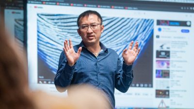 "Book of Mountains and Seas" composer Huang Ruo, a middle aged Asian man in a blue button down shirt, stands in front of a screen and discusses his creative process during a Virginia Tech class visit.