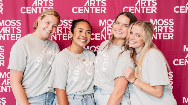 Members of a cappella group Soulstice pose in front of a maroon step and repeat banner featuring the Moss Arts Center repeated in white. Three young white women and one young Asian woman wear neutral Soulstice T-shirts with jeans.