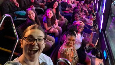 Virginia Tech senior and Moss Arts Center student employee Rhea Perdue (bottom left) gives a RideNYC tour. Perdue is a white woman with brown hair pulled back into a bun wearing black-framed glasses. She smiles widely, a full tour bus visible over her shoulder.