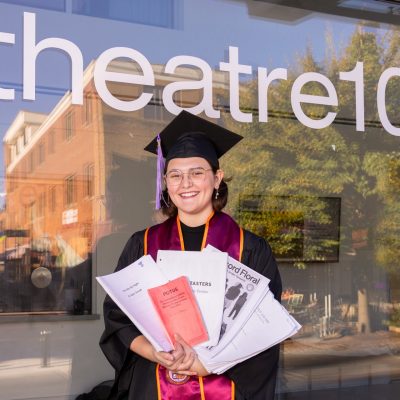 Virginia Tech senior and Moss Arts Center student employee Rhea Perdue holds scripts for the various plays she has participated in over the course of her time at the university. Perdue is a young white woman with short dark brown hair and glasses wearing a graduation cap and gown in front of the glass wall of Theatre 101 on Virginia Tech's Blacksburg campus.