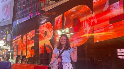 Virginia Tech senior and Moss Arts Center student employee Rhea Perdue in front of a Ride NYC tour bus, for which she hosts tours. Perdue is a young white woman with long, wavy dark brown hair wearing a light blue T-shirt and floral skirt.