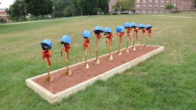 Ten gold shovels, each with a maroon and orange ribbon tied around the handle and a blue hard hat propped on top, stand upright in a dirt pit ahead of the official groundbreaking ceremony at the site of the Moss Arts Center.