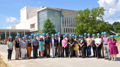 Faculty and staff for the Moss Arts Center and the Institute for Creativity, Arts, and Technology stand out front of the Moss Arts Center as it nears completion. They all wear cerulean blue hard hats.