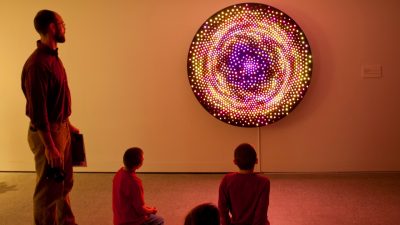 Patrons explore the galleries at the Moss Arts Center during opening week events. A middle aged man and two young boys view an LED art piece by Leo Villareal, a circular work with multicolored pink, purple, orange, and yellow lights.