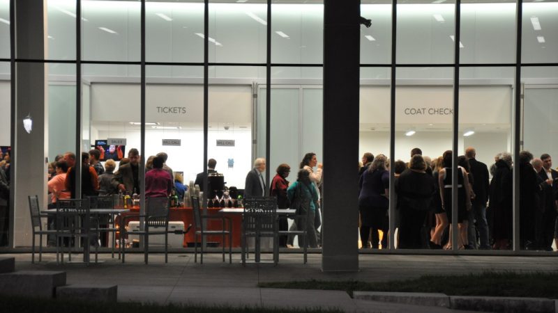 Image taken from the Moss Arts Center's front patio looking into the lobby, at evening. The lobby is well lit and full of guests before a performance. The Box Office and Coat Check stations are busy with guests.