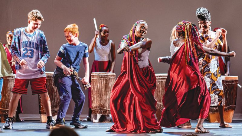 Area students in grades seven through 12 (at left) dance on stage with members of Ingoma Nshya, the Women Drummers of Rwanda, and playwright Kiki Katese (far right)