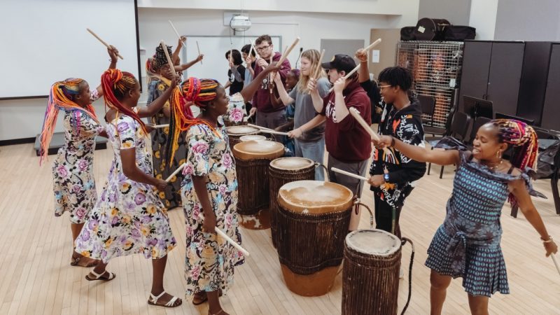 Members of Ingoma Nshya, the Women Drummers of Rwanda, play large African drums with Virginia Tech students during a class visit. The drummers are Black women in colorful, patterned dresses with rainbow colored braids pulled back into ponytails.