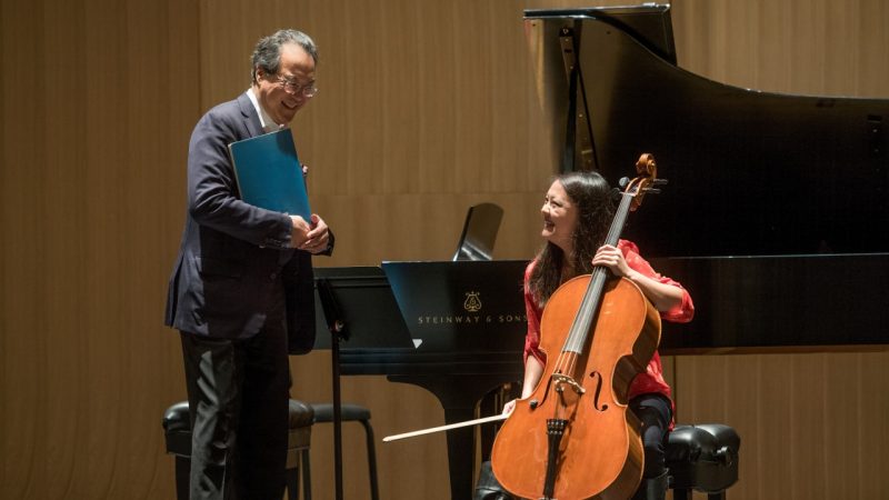  Cellist Yo-Yo Ma, a middle aged Asian man with glasses and wearing dark pants and a dark blazer, holds a book of sheet music and smiles widely at a student cellist, an Asian woman with long dark hair, who smiles widely back at him. She sits on a bench in front of a grand piano, holding her cello between her knees.