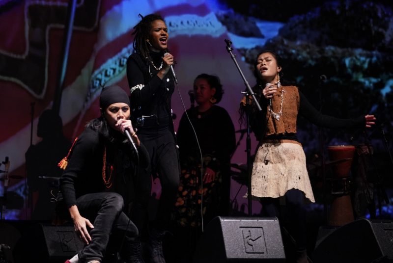 Three artists perform onstage singing. One person kneels, wearing a black jacked and headscarf, another stands with a black jacket on, and another standings singing with a brown top and cream skirt. Other musicians perform behind them and they all stand in front of a multi-colored, tapestry-looking projections.