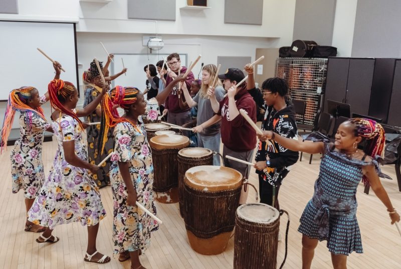 Women drummers from Rwanda teach their drumming techniques to a group of Virginia Tech students. The musicians and the students stand in the middle of a room, circling six big barrel drums, many of them holding drumsticks high in the air, ready to make contact with the drums. The drummers are are Black women with brightly colored long braids.  They wear multi-colored patterned dresses. The students are dressed in sweatshirts and jeans and sweatpants.