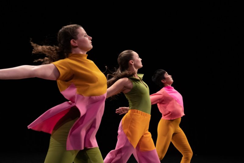 Three women dancers of Mark Morris Dance Group perform "The Look of Love" on stage, all facing towards the right edge of the frame, in front of a black background. The wear bright, color blocked uniforms.