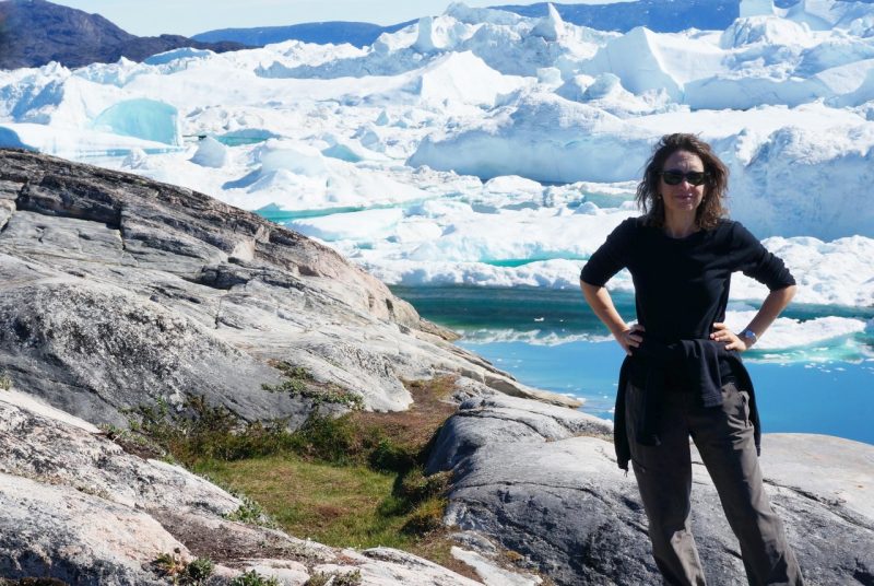 Science writer Elizabeth stands with her hands on her hips, posing in front of giant rocks and a glacier in blue-green water. She's wearing sunglasses, a black shirt and brown pants.