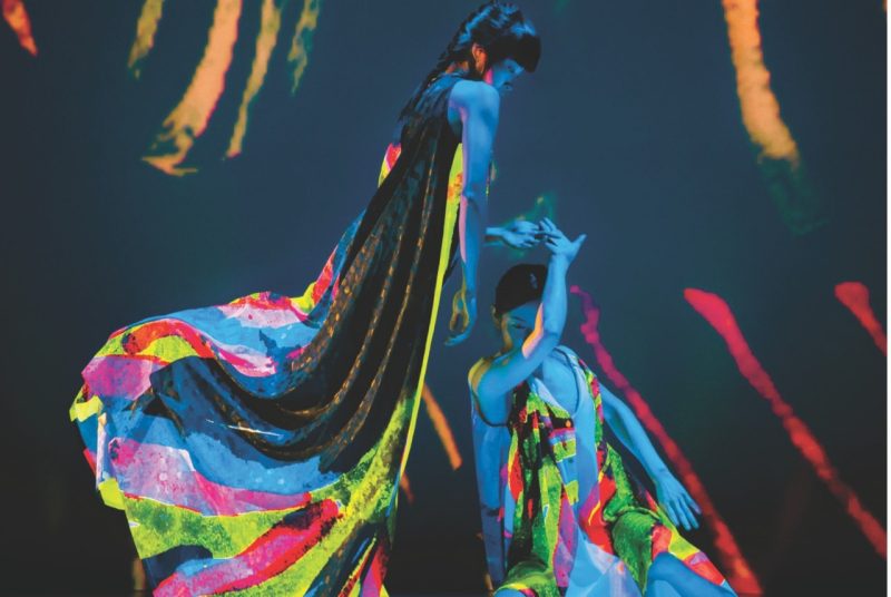 Two dancers with Cloud Gate Dance Theatre of Taiwan perform on stage wearing neon costumes that glows under a black light. One dancer sits while the other stands above her looking down. 