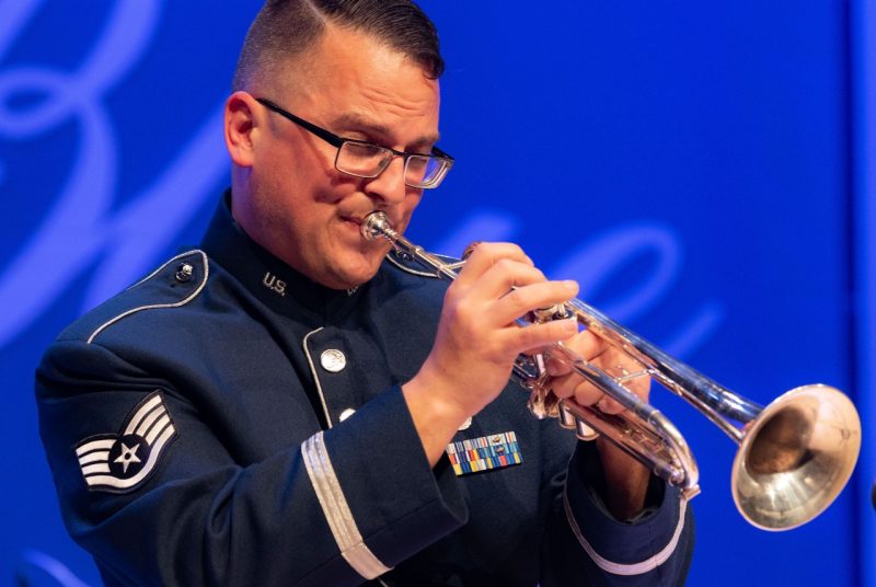 Staff Sgt. Nick Del Villano, a while man with short hair and glasses wearing a military uniform plays the trumpet onstage during a free concert at Delaware State University in Dover, Delaware.