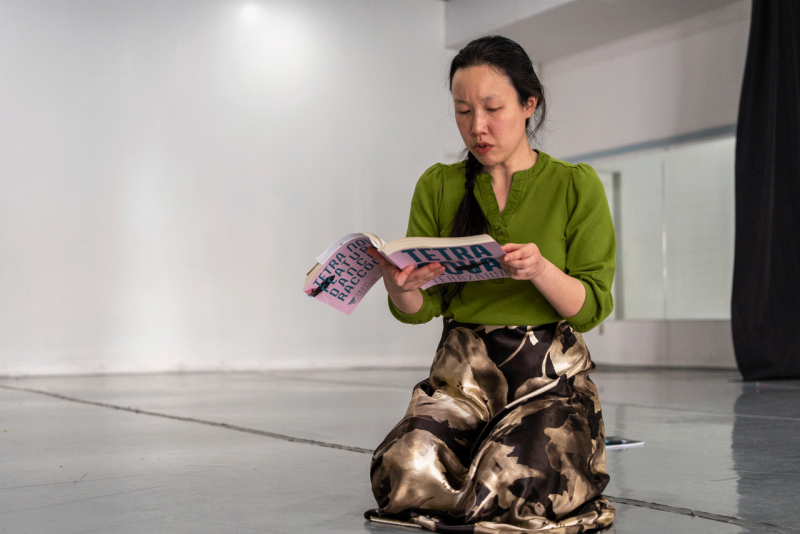 A woman kneels on the floor while reading her book, 'Tetra Nova.' She wears a green shirt and brown flowered shirt.