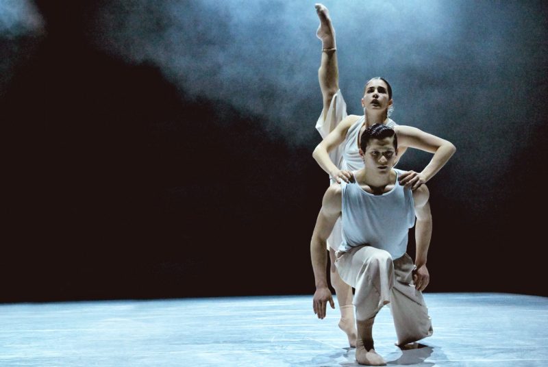 On a dimly lit stage, two contemporary dancers with Ballet BC, dressed in in white costumes, perform a duet. One kneels forward while the other balances behind with a leg extended straight upward.