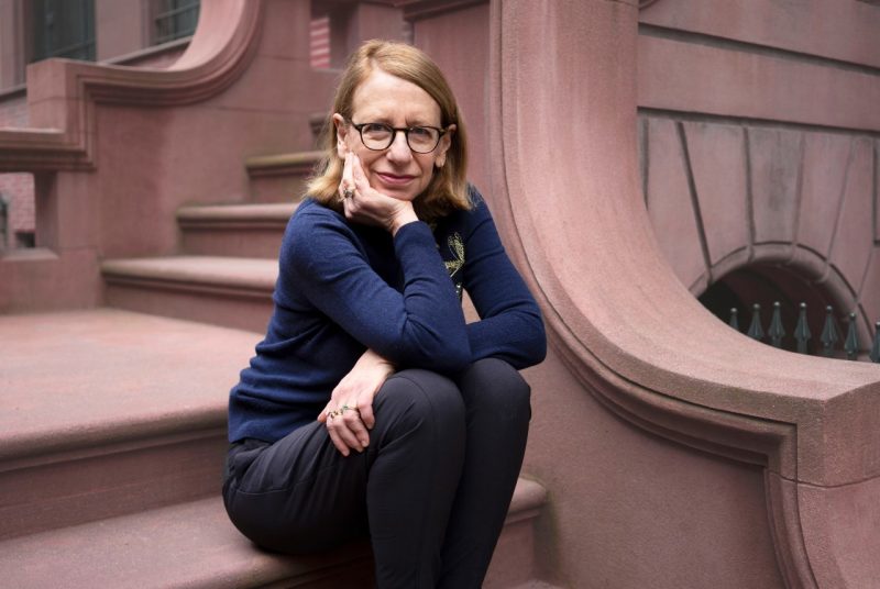 Roz Chast, a woman with short light brown hair and glasses, sits on the steps of a brownstone building, resting her chin on her hand and smiling slightly. She wears a navy sweater and dark pants.