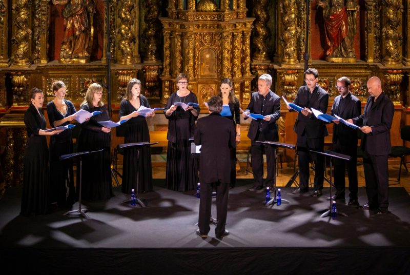 Ten male and female singers dressed entirely in black form a semi-circle and sing while each looks down at a book of music. A conductor in front facing the singers leads the group. The singers are in a church, standing in front an ornate, golden alter.