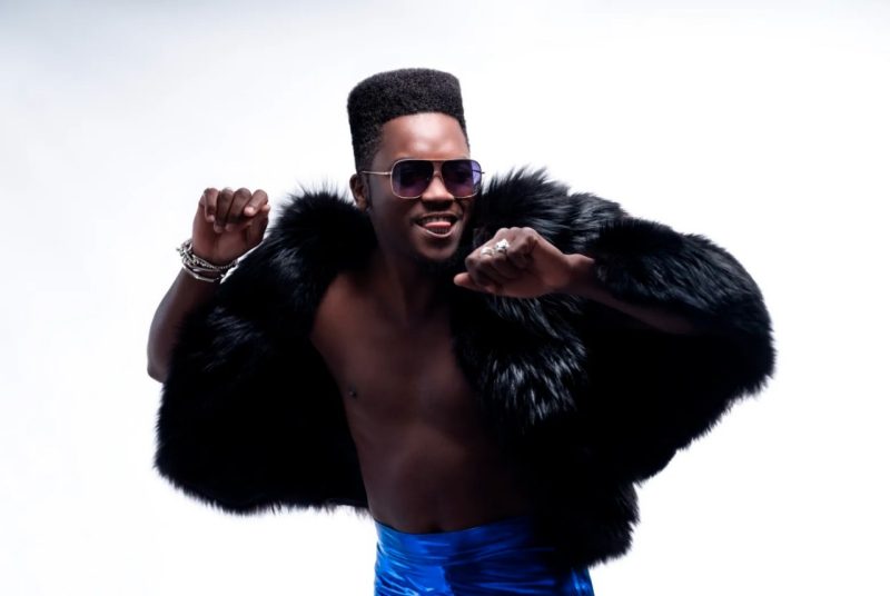 Musician Cimafunk poses shirtless in mid-dance, arms up and tongue out, wearing sunglasses, electric blue pants, and a furry jacket. 