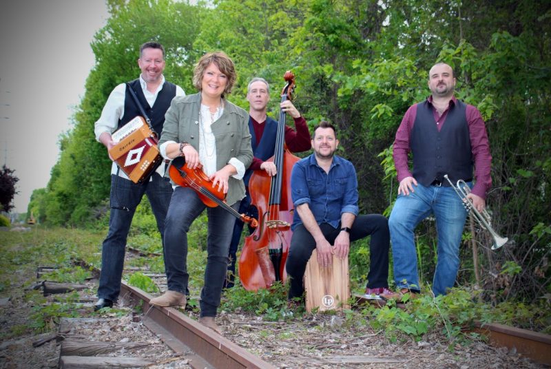 Eileen Ivers poses with her band, Universal Roots. The five musicians stand on an old train track, surrounded by trees and gravel. Eileen stands in front with her violin resting on her thigh, foot propped up on the train track. The four other male members pose closely behind her with their instruments. All are dressed casually, in jeans and long-sleeved shirts.