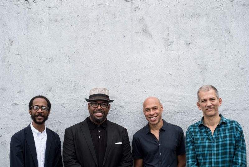 The four musicians who will perform "A MoodSwing Reunion" pose in front of a textured concrete wall. Drummer Brian Blade wears a white shirt with a black jacket, bassist Christian McBride wears a black shirt and jacket, black-rimmed glasses, and a beige hat, saxophonist Joshua Redman wears a black short-sleeved shirt, and pianist Brad Mehldau wears a green and black plaid button-up shirt.