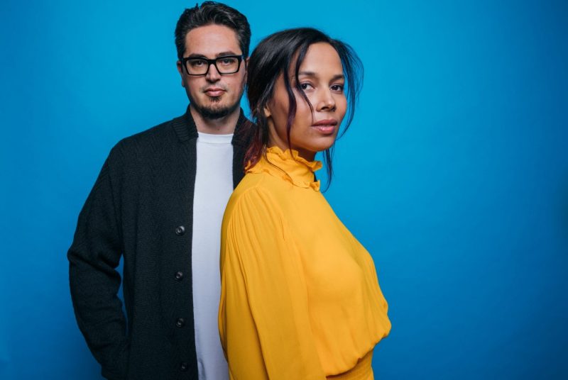 Musicians Rhiannon Giddens and Francesco Turrisi pose in front of a bright blue background. Rhiannon stands in front, wearing a mustard yellow dress, her body turned to the side while she looks into the camera. Francesco stands behind her, wearing dark-rimmed glasses, a white t-shirt and a dark casual jacket, staring directly at the camera.
