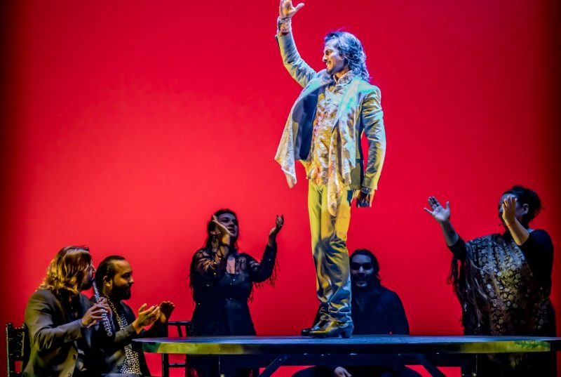 Flamenco dancer Farruquito stands on top of a table, hand reaching into the air, dressed in a shiny pearl-colored suit. Five people are around the table, some playing instruments, others clapping and cheering him on.