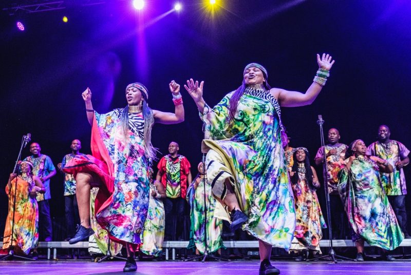 Two members of Soweto Gospel Choir dance at the front of a stage. They both have one foot off the ground and their hands in the air. They're wearing one-shouldered, flowing robes with beaded jewelry on their wrists and around their necks. Behind them, the rest of the choir sings in colorful attire.