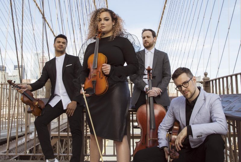 Four members of PUBLIQuartet pose with their instruments on a bridge. A female stands in front in a black dress, behind her are three men in blazers and button-up shirts.
