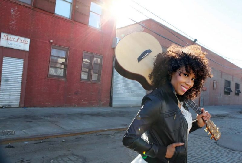 Country musician Rissi Palmer, a light-skinned Black woman with curly brown hair, wears a black leather jacket and a white shirt, and stands in the street in front of run down red brick commercial buildings. She holds an acoustic guitar by the neck and props the body up on her left shoulder.