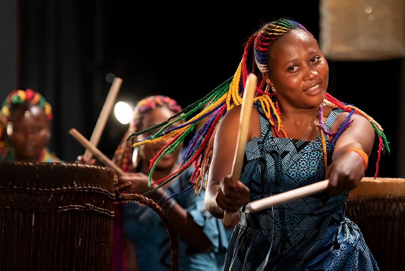 A Rwandan drummer performs "The Book of Life" on stage. She is a young Black woman with long, brightly colored braids, and she wears a blue and brown patterned dress.